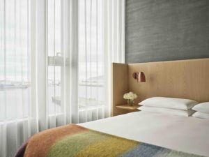 A queen bed with adjustable reading lamp next to a window with light-filtering drapes