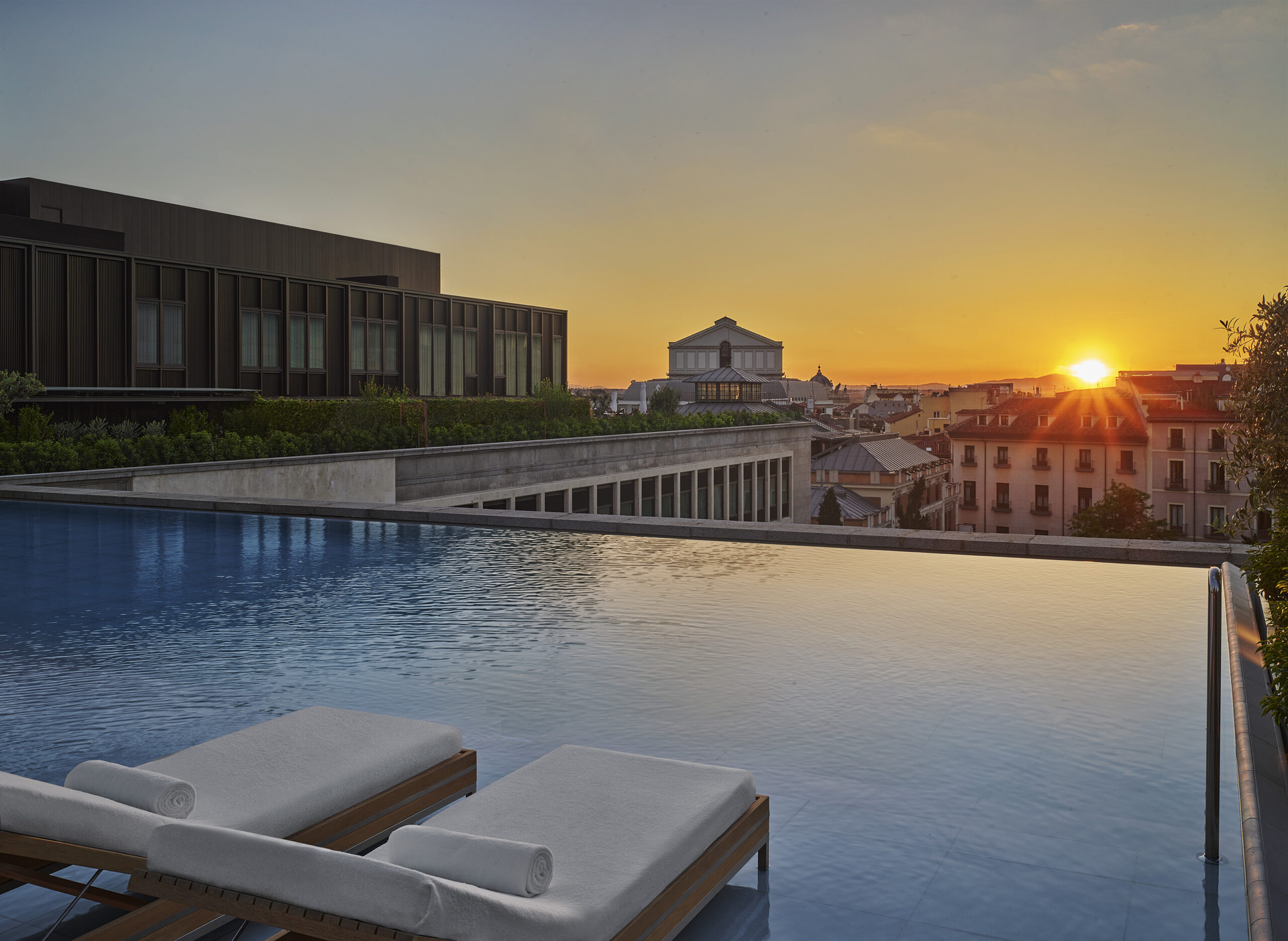Lounge chairs set inside an outdoor pool with a view of the sun setting over Madrid.